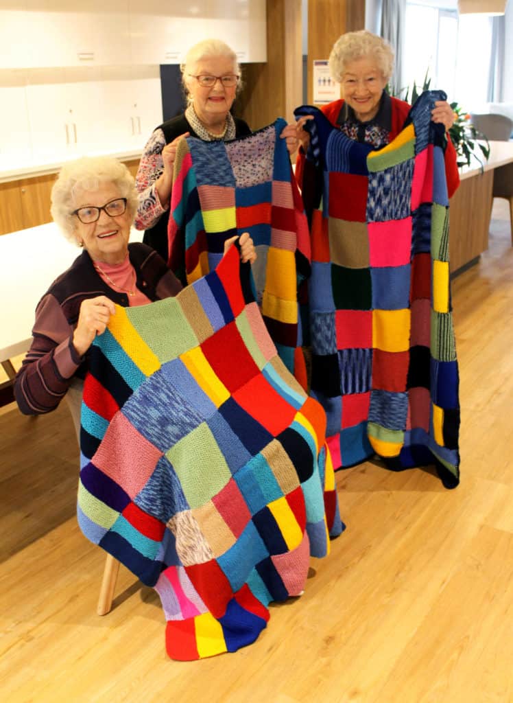 Three older ladies holding up colourful hand knitted blankets