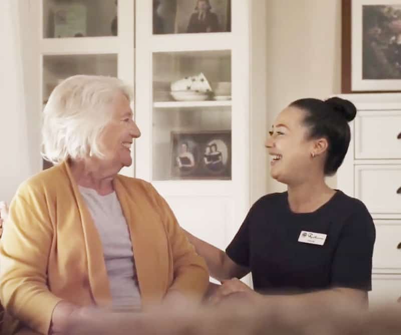 Older lady with white hair in a yellow cardigan smiling and talking with a young lady with dark hair in a bun and a black tshirt with a name tag.