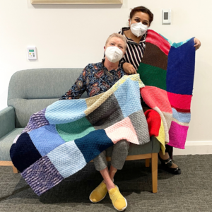 Two women seated on a lounge displaying knitted blankets