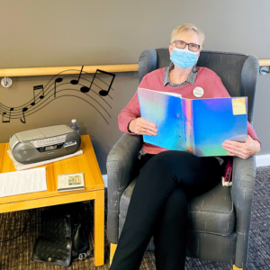 Volunteer Carolyn sits in a chair holding a blue folder open. She has a mask and is listening to the tape player on the coffee table next to her.