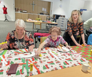 Elderly women doing Christmas craft with assistance from female volunteer in patterned blouse