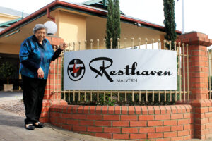 Elderly woman in blue jumper standing outside Resthaven Malvern sign