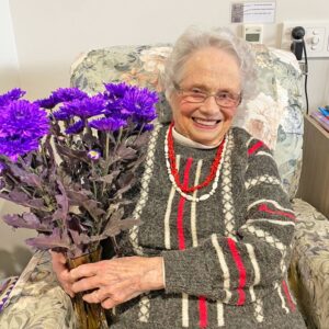 Mrs Audrey Niesche sitting in her armchair holding a bouquet of purple flowers