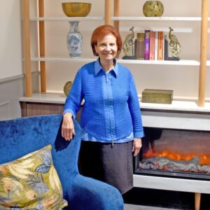 Maria Filpi wears a blue cardigan and stands inside Resthaven Leabrook next to the fireplace, smiling